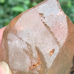 Può includere: Primo piano di un cristallo di quarzo traslucido color pesca. Il cristallo ha una superficie ruvida e strutturata con fratture e inclusioni interne. La luce cattura le sfaccettature, rivelando la sua forma naturale. Un esemplare minerale naturale.