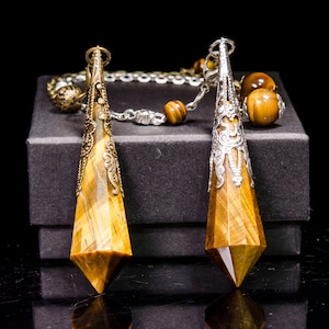 May include: Two gold-toned and silver-toned pendulums with tiger's eye stones hanging from a chain with tiger's eye beads. The pendulums are on a black velvet box.
