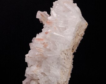 May include: A large, white crystal cluster with pink accents sits on a wooden base. The crystal is translucent and has a rough, natural texture.