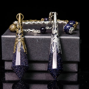 May include: Two blue and gold pendulums with intricate metalwork designs hang from a chain. The pendulums are displayed on a black velvet box.