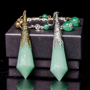 May include: Two green crystal pendulums with ornate silver and gold metal accents. The pendulums are hanging from a chain and are resting on a black box.