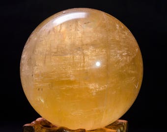 May include: A large, golden yellow crystal ball sits on a wooden stand. The ball is smooth and polished, with a slight sheen.