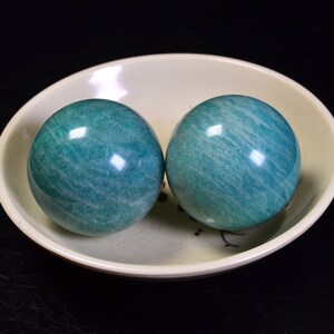 May include: Two green jade spheres, also known as Baoding balls, are resting in a white ceramic bowl. The balls have a smooth, polished surface with visible striations.