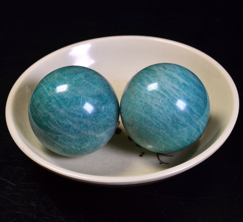 May include: Two green jade spheres, also known as Baoding balls, are resting in a white ceramic bowl. The balls have a smooth, polished surface with visible striations.