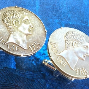 Op de afbeelding: Een paar zilverkleurige manchetknopen met een muntachtig ontwerp met een gedetailleerd profiel van een man en de inscriptie "IMP CAE SAR". De achtergrond is een blauwe stof met textuur.