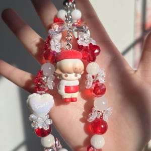 May include: A handmade charm bracelet featuring red, white, and clear beads. The bracelet includes a small figurine wearing a red beret and a white and red outfit. The charm is attached to a silver chain.