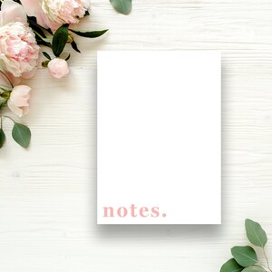Puede incluir: Un bloc de notas blanco en blanco con la palabra "notes." impresa en rosa sobre un fondo de madera blanco con flores rosas y hojas verdes.