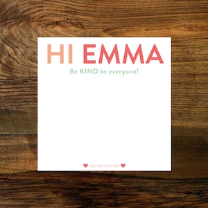 May include: White card with the text "HI EMMA" in pink and red letters. The text "Be KIND to everyone!" is written in green below the name. The bottom of the card has two red hearts and the text "you are so loved".