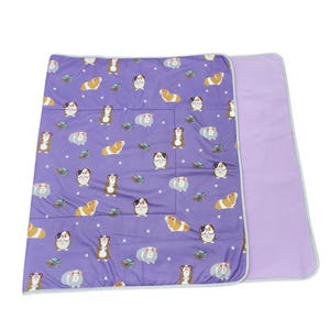 May include: A rectangular purple blanket featuring a repeating pattern of cartoon guinea pigs, some wearing glasses, interspersed with small stacks of books and white stars. The blanket has a solid light purple underside and light gray trim.