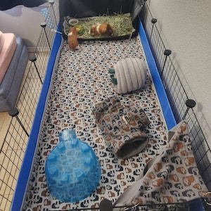 May include: A rectangular guinea pig cage with black wire walls and a blue plastic base. The interior features a white fabric liner with a repeating guinea pig pattern, a blue plastic igloo, a gray fabric tunnel, a gray hideout, and a hammock.