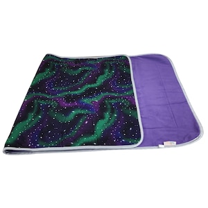 May include: A rectangular blanket featuring a dark purple and black background with a vibrant green and purple aurora borealis pattern and white stars. The reverse side is solid purple, and the entire blanket is finished with a light gray trim.