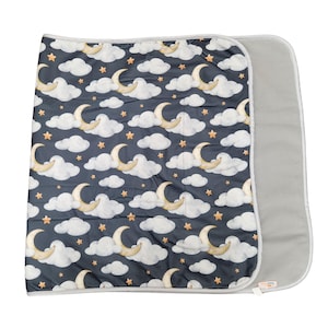 May include: A rectangular blanket featuring a dark navy blue background with a repeating pattern of watercolor-style white clouds, golden crescent moons, and small orange stars. The blanket has a solid light gray underside and gray trim.