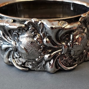 May include: Silver-toned metal coaster with an ornate, floral design. The coaster has a raised, circular center and a scalloped edge.