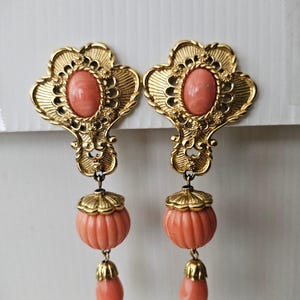 May include: A pair of vintage gold-tone earrings with coral-colored accents. Each earring features an ornate, filigree-style top with an oval coral-colored stone. Below, a coral-colored pumpkin-shaped bead hangs, finished with a coral-colored teardrop.