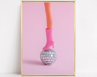 May include: A framed print featuring a doll's leg and a pink boot atop a disco ball. The doll's leg is peach-colored, and the boot is a vibrant pink. The disco ball is silver and reflective, set against a soft pink background.