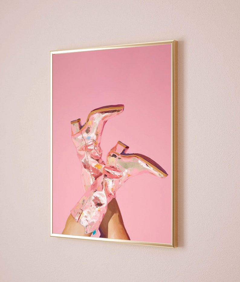 May include: A pair of pink cowboy boots with a metallic finish are propped up against a pink background. The boots are in focus and the background is blurred.