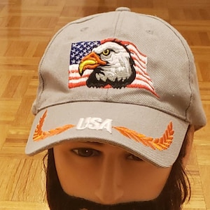 May include: A grey baseball cap with an embroidered American flag and eagle design. The word "USA" is embroidered on the brim, with orange feather designs. A patriotic accessory, suitable for casual wear.