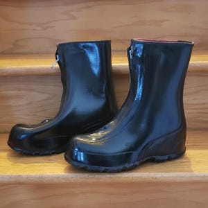 Peut inclure: Une paire de bottes en caoutchouc noires avec une finition brillante. Les bottes ont une fermeture éclair et un bout arrondi. Les bottes sont posées sur une surface en bois.