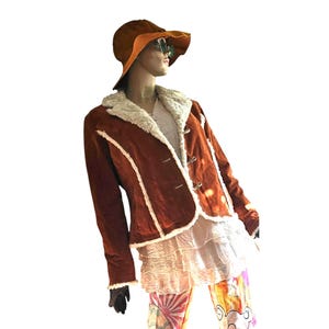 Peut inclure: Une veste couleur rouille avec une bordure en fausse fourrure blanche le long du col, de l'ouverture avant et des poignets. La veste présente des fermetures à bascule et est portée avec un chapeau à larges bords rouille et orange, un haut blanc à volants et un pantalon à motifs graphiques.