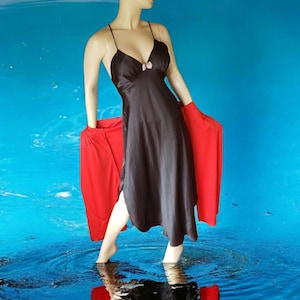 May include: A black satin slip dress with spaghetti straps and a small bow detail at the bust. A red shawl is draped over the mannequin's arms. The mannequin is standing in a blue water-like background.