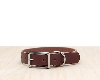 May include: A brown leather dog collar with a silver buckle and D-ring. The collar is made from a single strip of leather and has adjustment holes. The collar is displayed on a light wood surface.