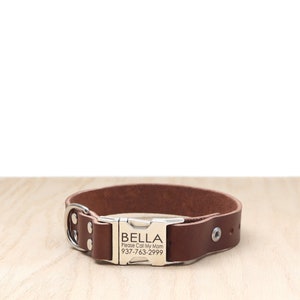 Puede incluir: Collar de perro de cuero marrón con una hebilla plateada. La hebilla tiene el nombre "BELLA" grabado, junto con el texto "Please Call My Mom" y un número de teléfono.