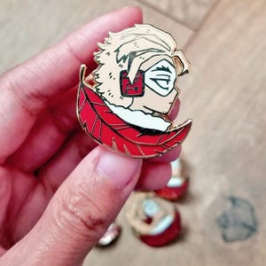 Puede incluir: Un pin de esmalte con bordes de metal dorado que muestra un personaje estilizado con cabello rubio puntiagudo, una máscara blanca y auriculares rojos. El personaje se sitúa sobre una gran pluma roja curvada con detalles en negro.