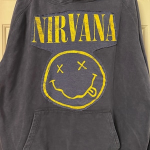 May include: Dark gray hoodie featuring the word "NIRVANA" in yellow block letters above a yellow smiley face with crossed-out eyes and a protruding tongue. The hoodie has a front pocket and a drawstring hood.