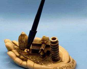 Könnte beinhalten: Eine weiße Keramikhand hält einen Stift, eine Miniatur-Nachbildung des Schiefen Turms von Pisa und anderer Gebäude. Die Hand ist mit dem Text "SOUVENIR di PISA" beschriftet.