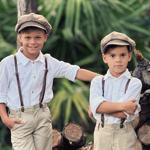 Puede incluir: Dos niños pequeños con camisas blancas, tirantes marrones y pantalones cortos beige se paran frente a una pila de troncos. Ambos niños llevan gorras de repartidor marrones.
