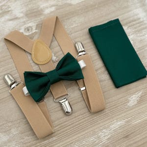 May include: A formal accessory set featuring tan suspenders with silver-tone clips, a dark green bow tie, and a matching pocket square. The bow tie and pocket square provide a pop of color against the neutral suspenders.