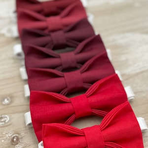 May include: A set of six bow ties in shades of red, ranging from a deep burgundy to a bright red. The bow ties are arranged in a line, with the darkest shade at the top and the lightest shade at the bottom.