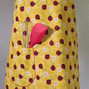 May include: Yellow apron with a ladybug and daisy pattern. The apron has a pocket with a whisk and red spatula. The bottom edge has a white lace trim. The straps are red with white polka dots.