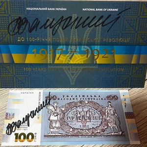 May include: A Ukrainian 100 Hryvnia banknote and commemorative box. The box is blue with gold accents and text in Ukrainian and English, celebrating the 100th anniversary of the Ukrainian Revolution. The banknote features historical figures.