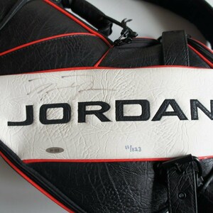 Puede incluir: Bolsa de golf en blanco y negro con el nombre "JORDAN" en letras negras grandes. La bolsa tiene ribetes rojos y un panel central blanco. Incluye un número de serie y el número "11/123".