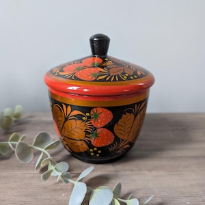 May include: A hand-painted wooden container with a lid. The container is black with orange, red, and gold floral and strawberry designs. The lid has a black knob. The container sits on a wooden surface.