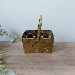 May include: A small, square, woven brass basket with a handle. The basket has a textured, interwoven design and a hinged handle. The brass has a warm, golden hue, and the basket is sitting on a wooden surface.