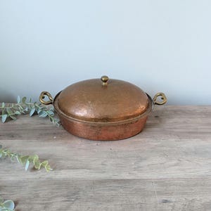 May include: A copper-colored, round lidded casserole dish with brass handles and a brass knob on top. The dish is sitting on a wooden surface. The lid has a hammered texture.