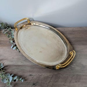 May include: Oval silver serving tray with gold-colored handles. The tray has a polished surface and a slightly worn appearance. The handles are curved and add a decorative element to the tray. A classic piece for serving food or displaying items.