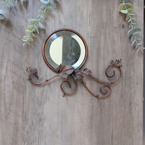 May include: A round, copper-toned mirror with a decorative metal vine and leaf design. The mirror is mounted on a wooden surface, with a simple, elegant aesthetic. The decorative metalwork features curling tendrils and stylized leaves.