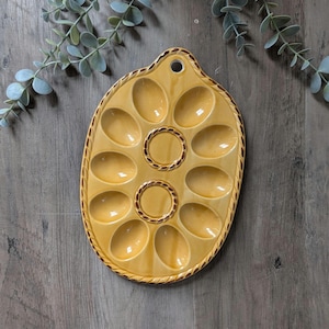 May include: Yellow ceramic escargot dish with 12 oval compartments and a decorative rope-like border. The dish has two circular indentations in the center and a small hole at the top for hanging. The background is a wooden surface.