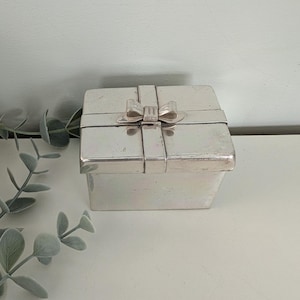 May include: A silver-colored, square box with a bow on top, resembling a gift. The box has a lid and is made of a shiny, metallic material. The bow is also silver and adds a decorative touch. The box is sitting on a white surface.