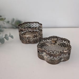 May include: Two antique silver-toned filigree boxes. One is a small, openwork cylinder. The other is a four-lobed box with a matching filigree design and a hinged lid. The lid is topped with a small floral accent.