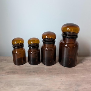 May include: Four amber glass apothecary jars with rounded lids. The jars are arranged in a row, varying in size from small to large. The lids are a lighter amber color than the jars. The jars are sitting on a wooden surface.