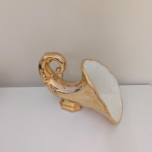 May include: A cornucopia-shaped vessel featuring a metallic gold exterior with a visible crackle finish and a smooth white interior. The object is supported by a small, rectangular gold base and displays a curved, horn-like silhouette.