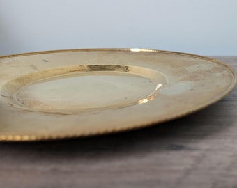 May include: A gold-colored plate with a beaded rim. The plate has a slightly concave center and a smooth, reflective surface. The plate is set on a wooden surface.