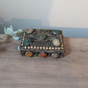 May include: A rectangular metal box featuring intricate silver-toned filigree wirework and a border of small metallic spheres. The lid and front panel are inlaid with several smooth, polished, multi-colored cabochon stones in shades of green, tan, and orange.