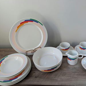 May include: White ceramic dinnerware set with rainbow-colored stripes. Includes plates, bowls, cups, saucers, and a creamer. The set features a clean, modern design, perfect for everyday use or special occasions. A complete dining set.