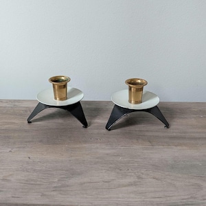 May include: Two mid-century modern candle holders. Each has a white disc, a brass cup, and three black legs. The candle holders are on a wooden surface.