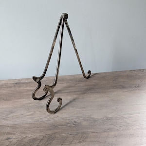 May include: A silver-toned metal display stand featuring a hinged, tripod-style design with two front-facing curved hooks for holding items and a single rear support leg. The stand has a smooth, reflective finish and a simple, functional structure.